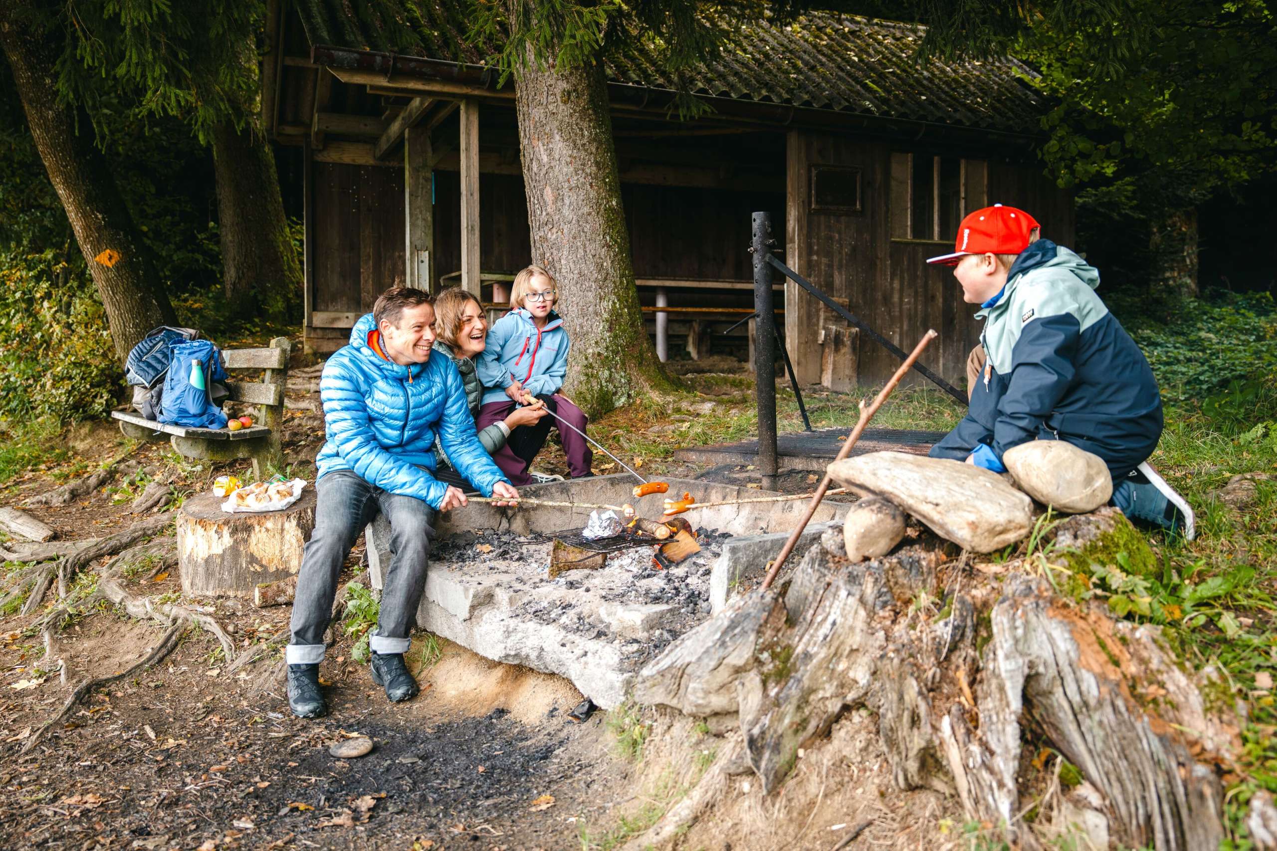 Familie mit 2 Kinder um den Grill am Waldrand stehend, sitzend Cervelat auf Stöcken grillend. Hütte und Bank mit Rucksack im Hintergrund.