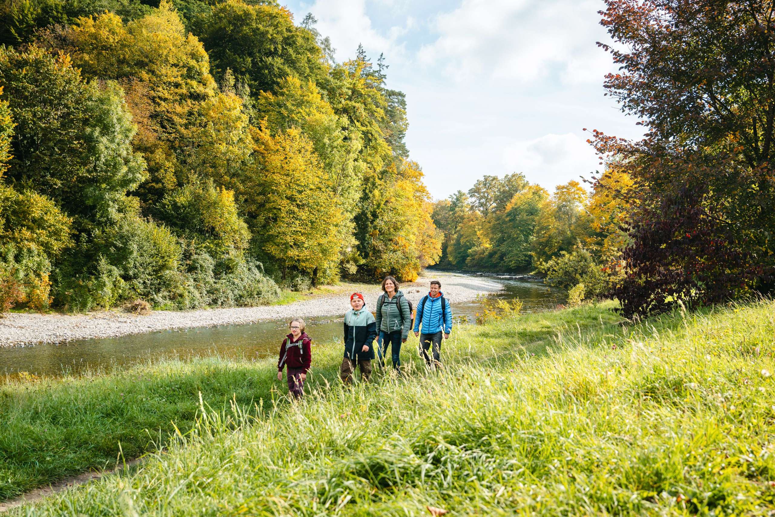 Spaziergang einer 4-köpfigen Familie in Burgdorf Nähe Waldeggbrücke entlang der Emme auf dem Trampelpfad, Uferweg zwischen Gras. Emme mit Steinen und Bäumen herbstlich in der Sonne leuchtend.